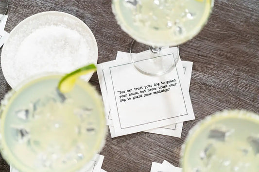 Organic cotton cocktail napkin printed with the quote "You can trust your dog to guard your house, but never trust your dog to guard your sandwich," styled with margarita glasses and a bowl of salt on a wood table, from Five Star Napkins' Margarita quote collection