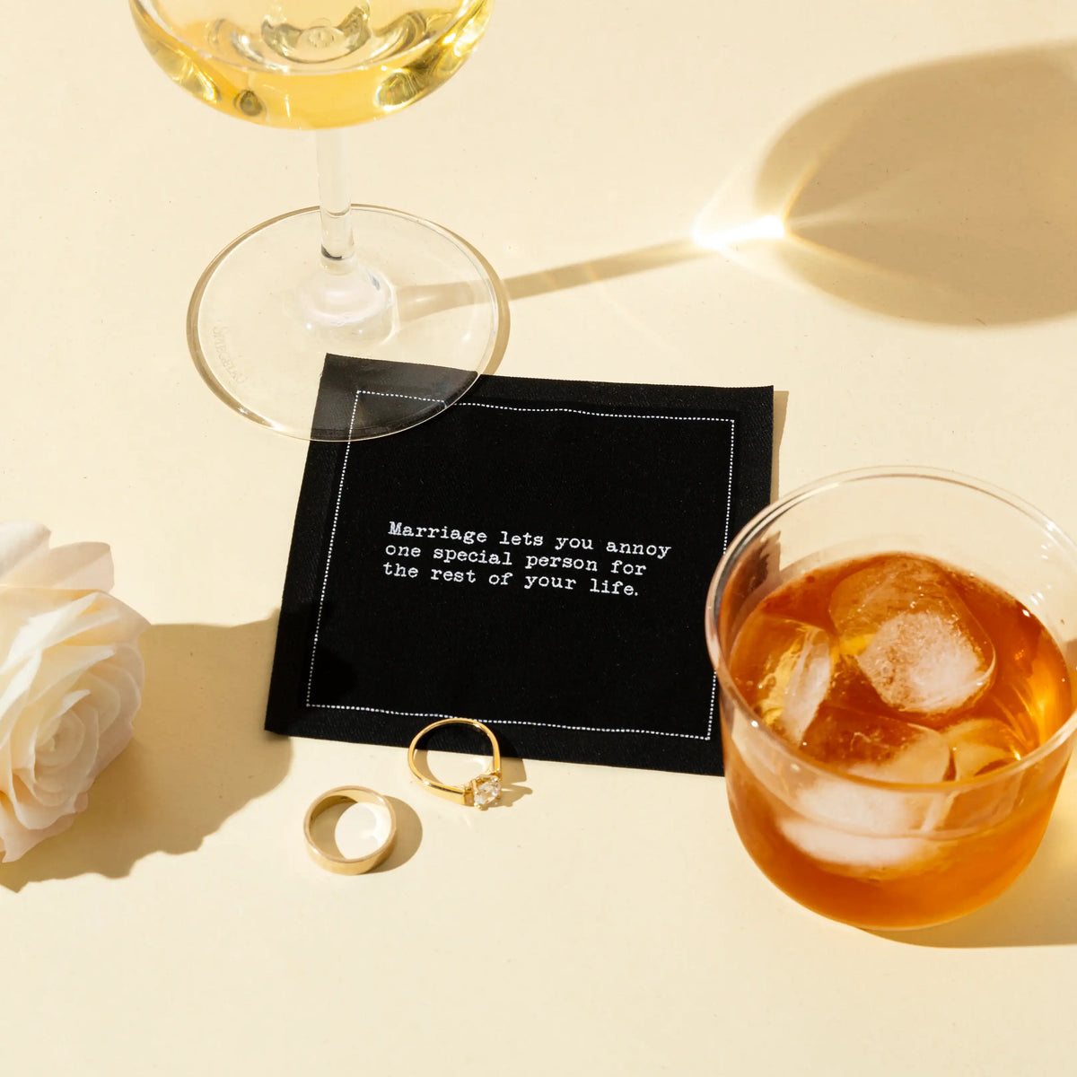 Black marriage quote cocktail napkin with white typewriter-style text reading, “Marriage lets you annoy one special person for the rest of your life,” styled with drinks, wedding rings, and a white rose.