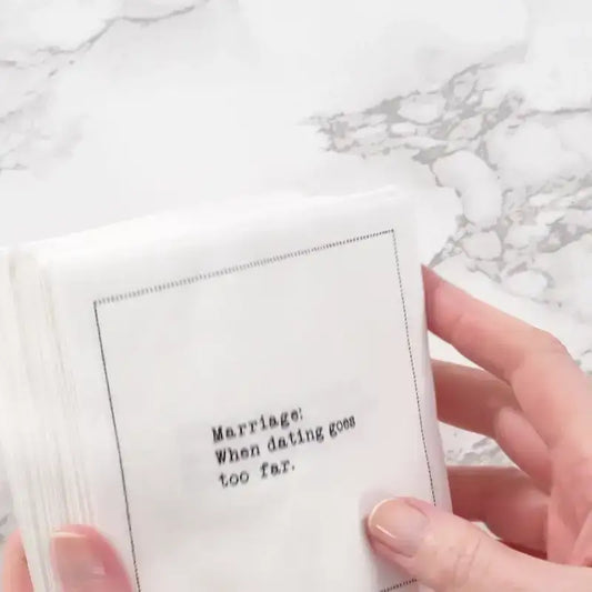 Hand holding a stack of white marriage quote cocktail napkins with black typewriter-style text reading, “Marriage: When dating goes too far.”