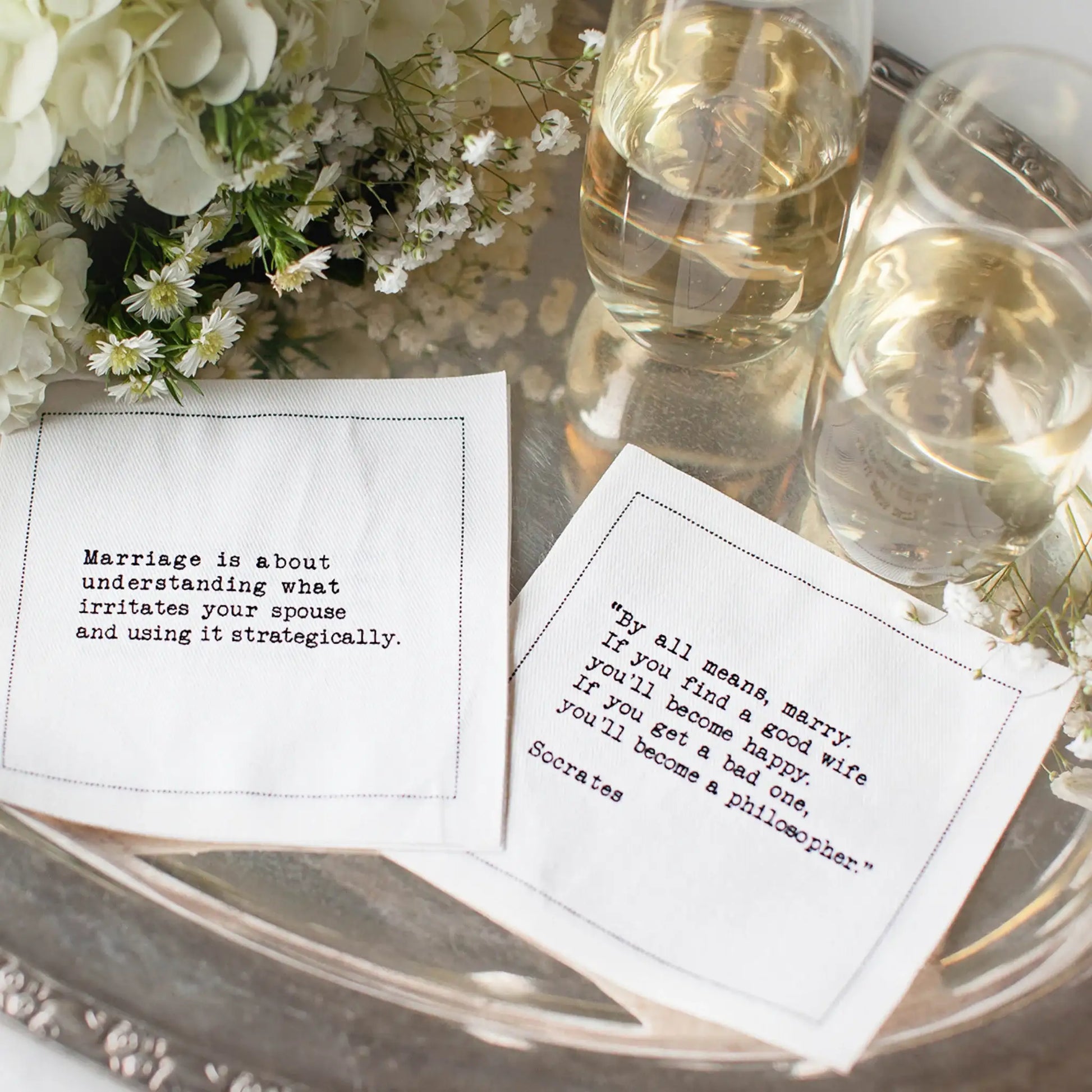 White marriage quote cocktail napkins with black typewriter-style sayings about married life, styled on a silver tray with white flowers and glasses of wine.