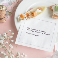 White marriage quote cocktail napkin with black typewriter-style text reading, “The secret of a happy marriage remains a secret.” – Henny Youngman, styled with appetizers, flowers, and a champagne cork.