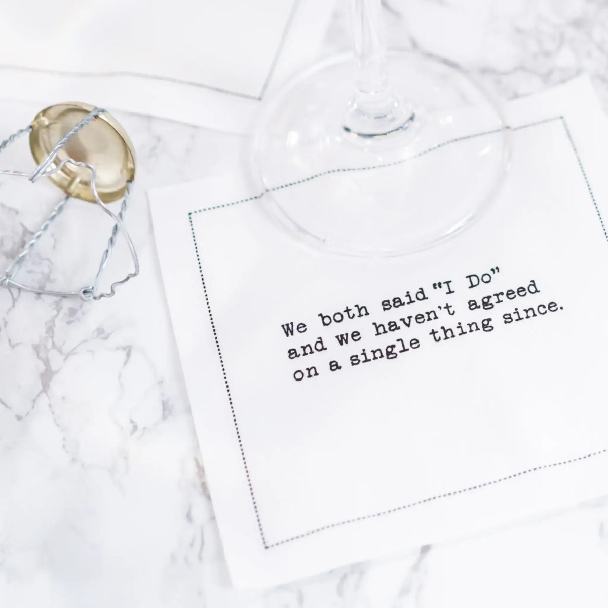 White marriage quote cocktail napkin with black typewriter-style text reading, “We both said ‘I Do’ and we haven’t agreed on a single thing since,” styled beneath a wine glass with a champagne cage nearby.