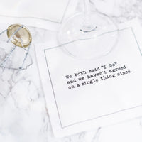 White marriage quote cocktail napkin with black typewriter-style text reading, “We both said ‘I Do’ and we haven’t agreed on a single thing since,” styled beneath a wine glass with a champagne cage nearby.