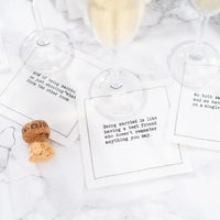 White marriage quote cocktail napkins with funny sayings about married life, styled beneath champagne glasses with a champagne cork on a marble surface.