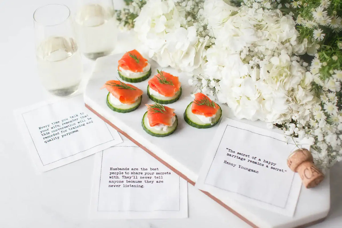 White marriage quote cocktail napkins with humorous sayings, styled alongside smoked salmon appetizers, white flowers, wine glasses, and a champagne cork.