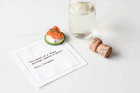 White marriage quote cocktail napkin with black typewriter-style text reading, “The secret of a happy marriage remains a secret.” – Henny Youngman, styled with a smoked salmon appetizer, wine glass, and champagne cork.