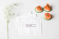 White marriage quote cocktail napkins with black typewriter-style text reading, “Never laugh at your wife’s choices. You are one of them...” styled with smoked salmon appetizers and delicate white flowers.