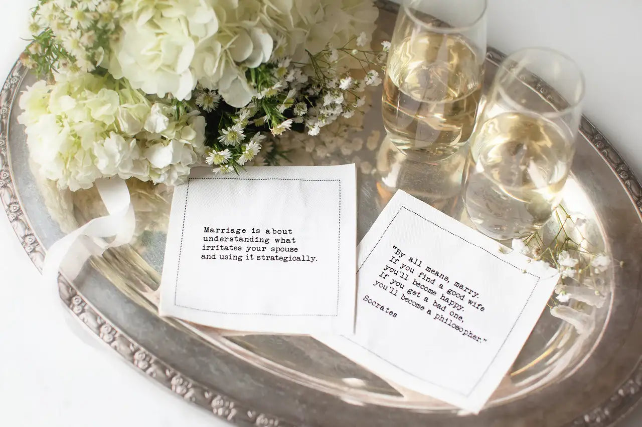 White marriage quote cocktail napkins with humorous sayings about married life, styled on a silver tray with white flowers and glasses of wine.