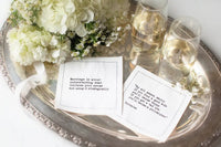 White marriage quote cocktail napkins with humorous sayings about married life, styled on a silver tray with white flowers and glasses of wine.