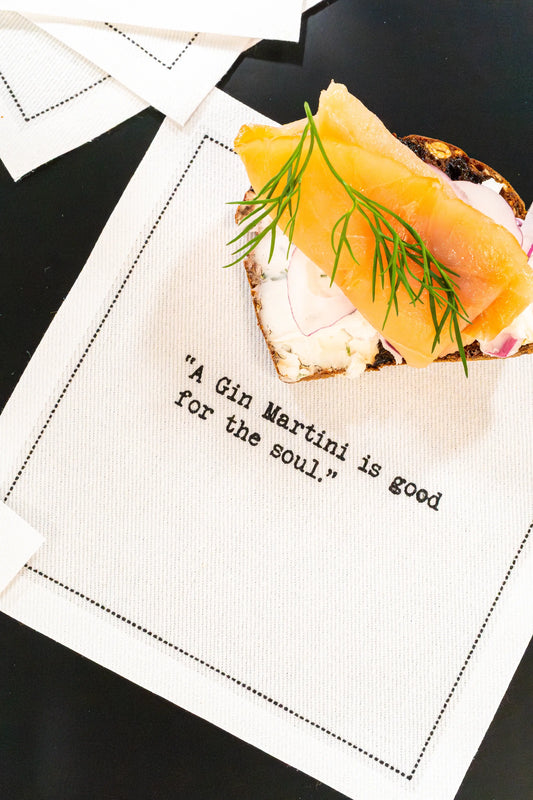 Organic cotton cocktail napkin printed with the quote "A Gin Martini is good for the soul," styled with an open-face smoked salmon toast, from Five Star Napkins' Martini quote collection