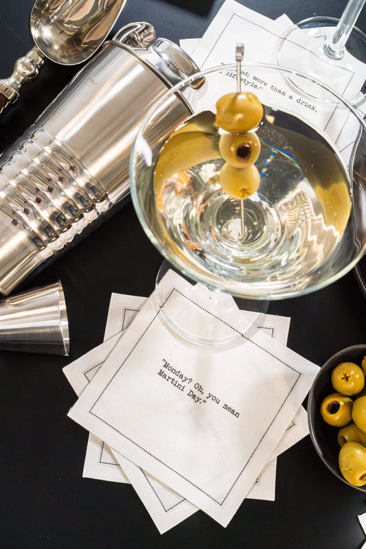 Organic cotton cocktail napkin printed with the quote "Monday? Oh, you mean Martini Day," styled with a martini glass, olives, and a cocktail shaker, from Five Star Napkins' Martini quote collection