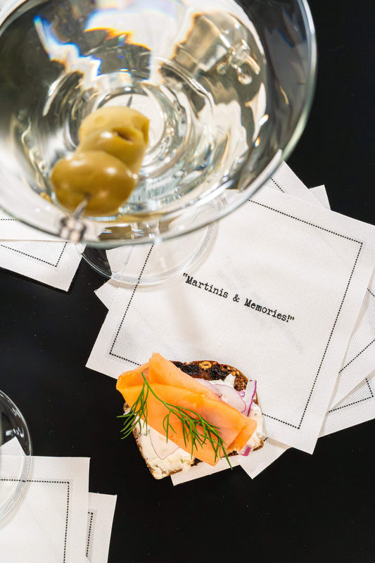 Organic cotton cocktail napkin printed with the quote "Martinis & Memories," styled with a martini glass and an open-face salmon toast, from Five Star Napkins' Martini quote collection