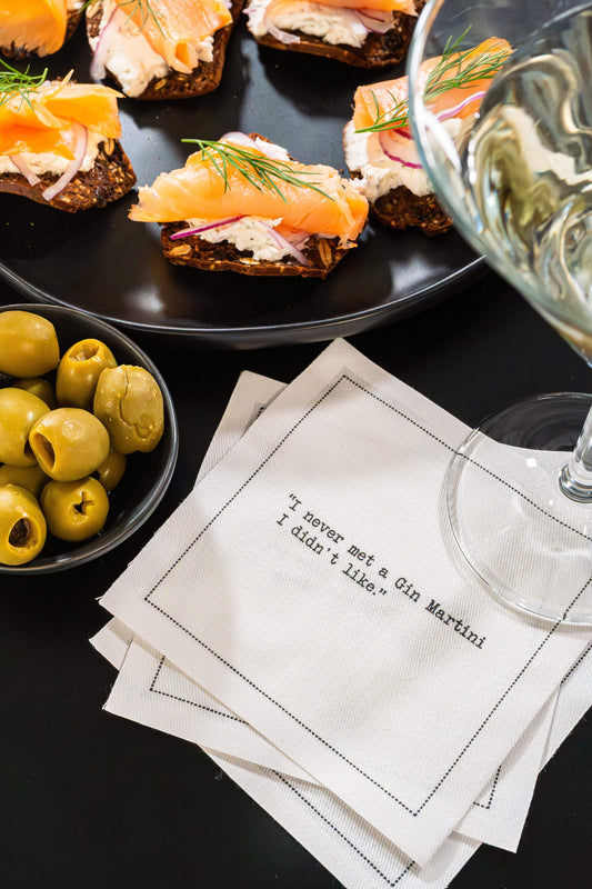 Organic cotton cocktail napkin printed with the quote "I never met a Gin Martini I didn't like," styled with olives, open-face salmon toasts, and a martini glass, from Five Star Napkins' Martini quote collection