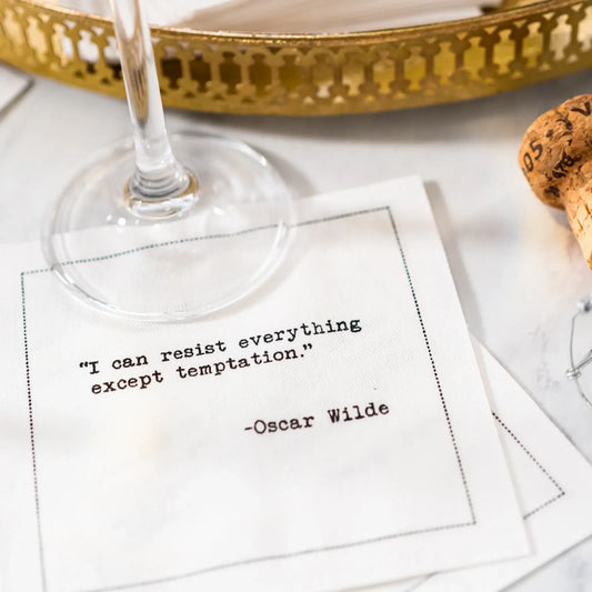 Organic cotton cocktail napkin printed with the quote "I can resist everything except temptation," styled on a marble table with a gold tray, wine glass, and champagne cork, from Five Star Napkins' Oscar Wilde quote collection