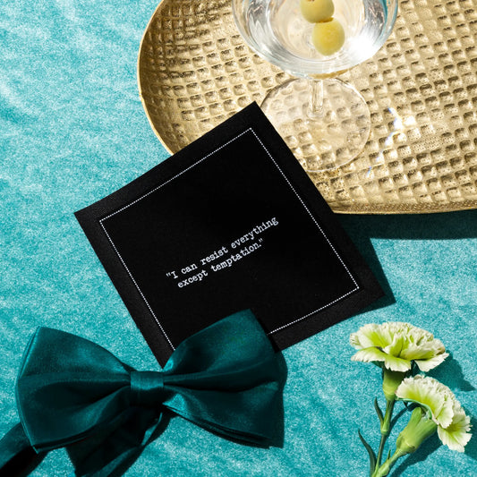 Organic cotton cocktail napkin, black, with white dotted border printed with the quote "I can resist everything except temptation," styled on a teal velvet surface with a martini glass, teal bow, and carnations, from Five Star Napkins' Oscar Wilde quote collection