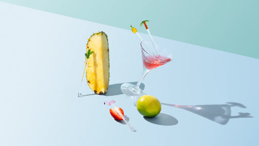 Glass swizzle sticks with pineapple and palm tree designs styled with a cocktail, pineapple, and lime, by Five Star Napkins.
