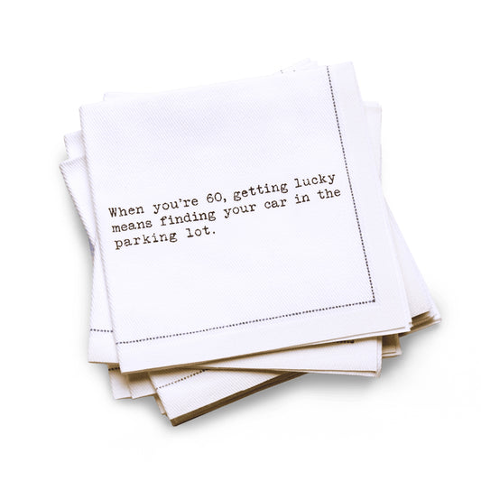 Organic cotton quarter-fold napkin with black dotted border printed with the quote "When you're 60, getting lucky means finding your car in the parking lot," from Five Star Napkins' 60th birthday quote collection
