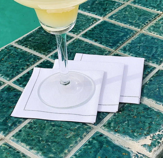 Three white organic cotton cocktail napkins folded on tiled table with a drink glass