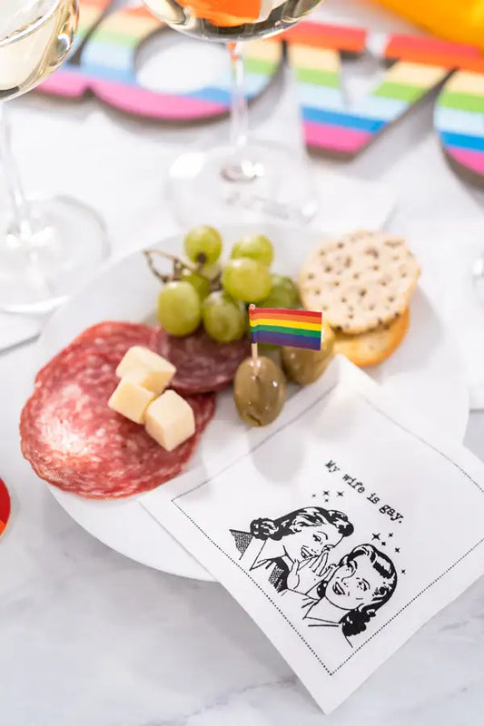 Organic cotton cocktail napkin with 'My wife is gay.' quote on a party plate with charcuterie and rainbow decor