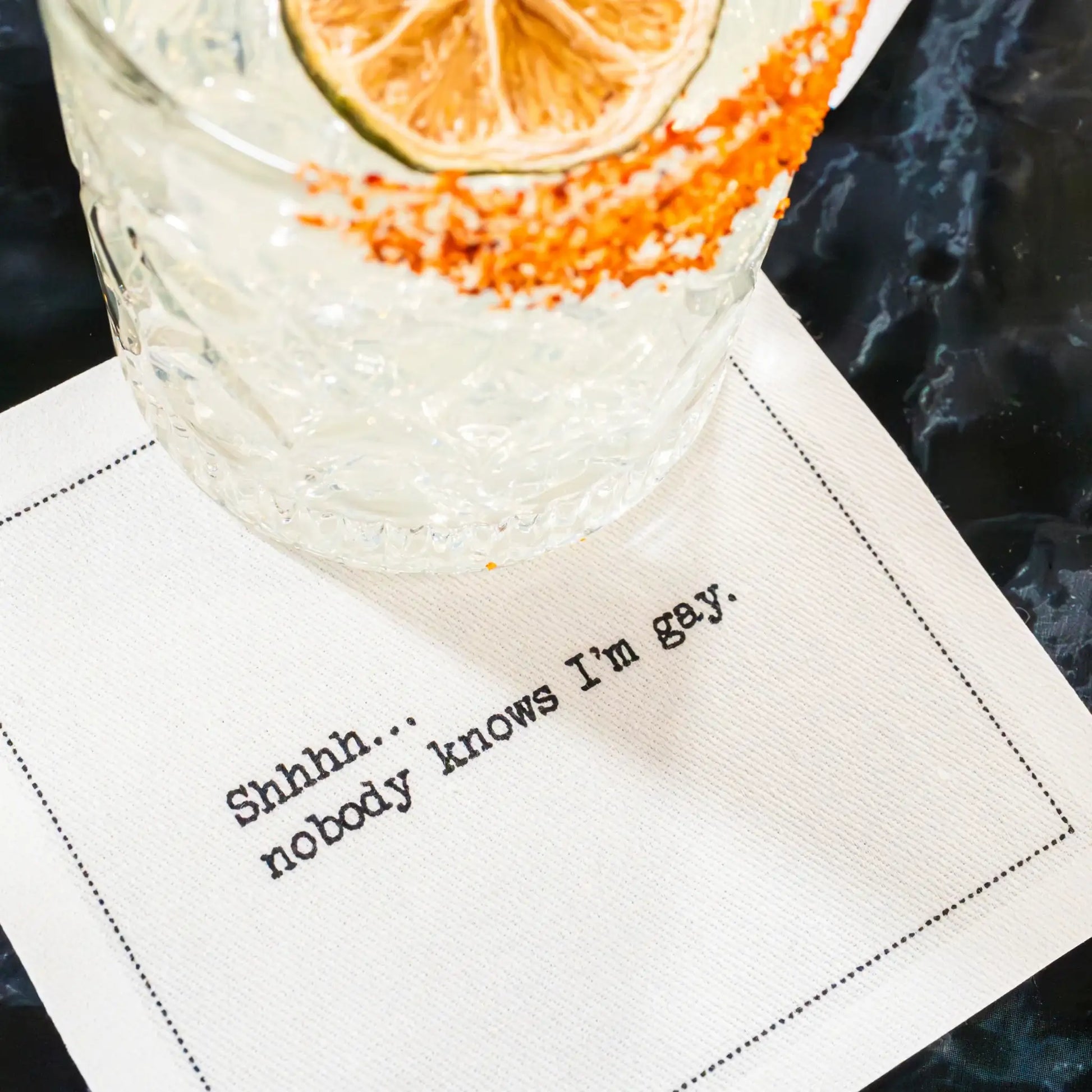 Organic cotton cocktail napkin with quote 'Shhhh... nobody knows I'm gay.' under a drink