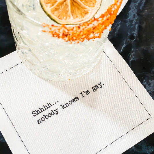 Organic cotton cocktail napkin with quote 'Shhhh... nobody knows I'm gay.' under a drink