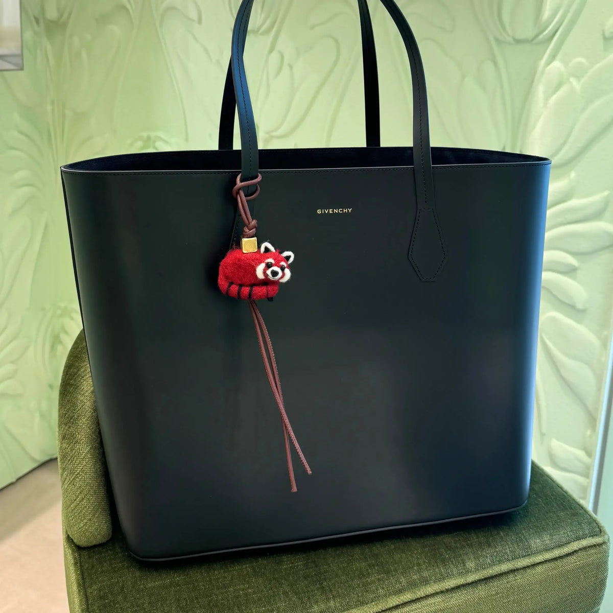 Black leather tote bag with a red panda felt keychain and brown leather strap, displayed on a green chair