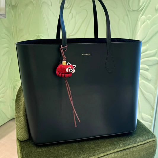 Black leather tote bag with a red panda felt keychain and brown leather strap, displayed on a green chair