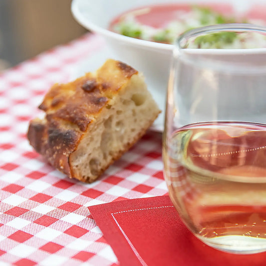 Glass of white wine, bread, and bowl on red checkered tablecloth with Persimmon organic cotton napkin