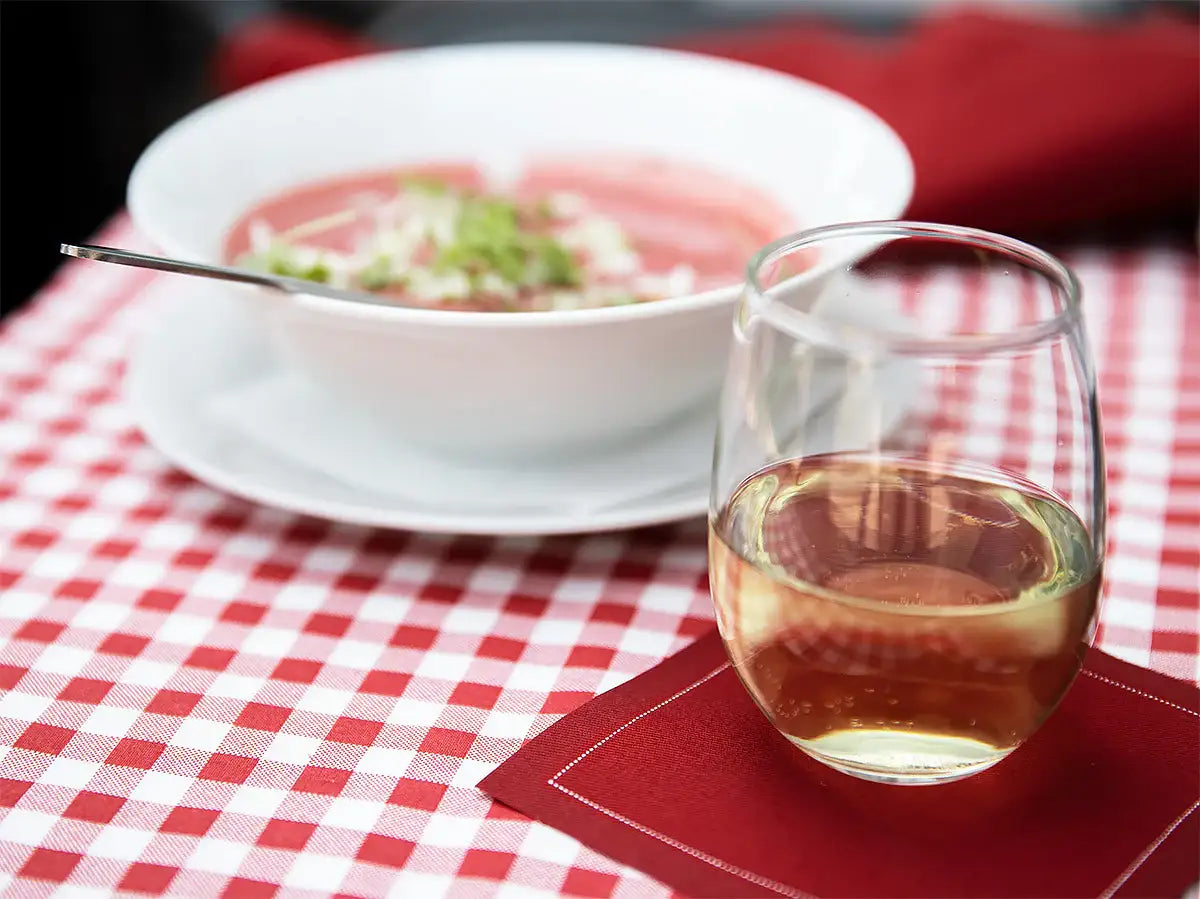 Glass of white wine on Persimmon organic cotton napkin beside bowl of soup on red check tablecloth