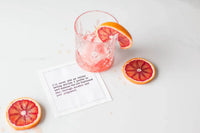 Organic cotton quote napkin with black printed text beside a cocktail glass and sliced citrus at a cocktail party.