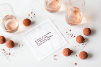 Organic cotton quote napkin with text 'My prince is not coming on a white horse. He's obviously riding a turtle and is lost,' styled with chocolate truffles and drinks on a white table.