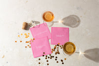 Pink cocktail napkins with playful wedding sayings are arranged beside two glasses of rosé, a champagne cork and cage, and scattered gold heart confetti.