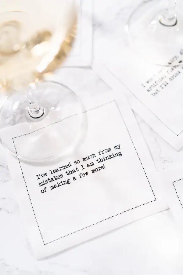 Organic cotton cocktail napkin printed with the quote "I've learned so much from my mistakes that I am thinking of making a few more!" styled under a wine glass on a marble table, from Five Star Napkins' Snarky quote collection