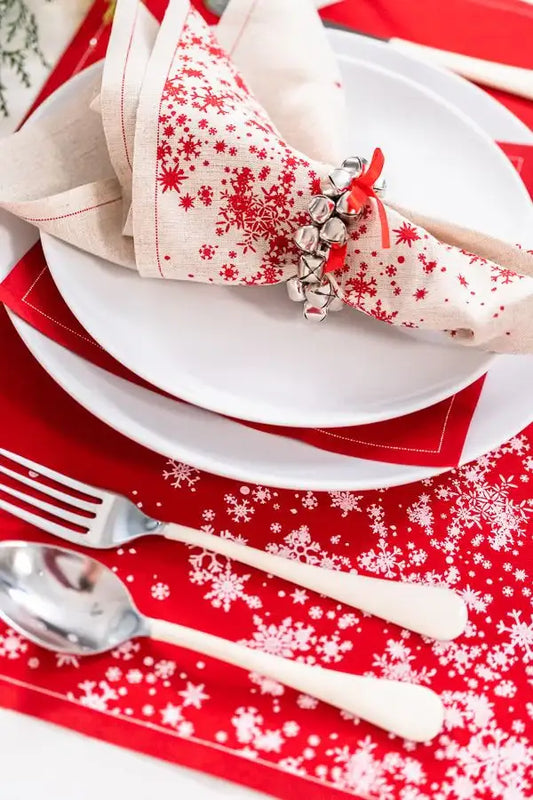 Red and natural organic cotton napkin with snowflake print folded on white plate, holiday table setting with matching placemat and silver jingle bell napkin ring