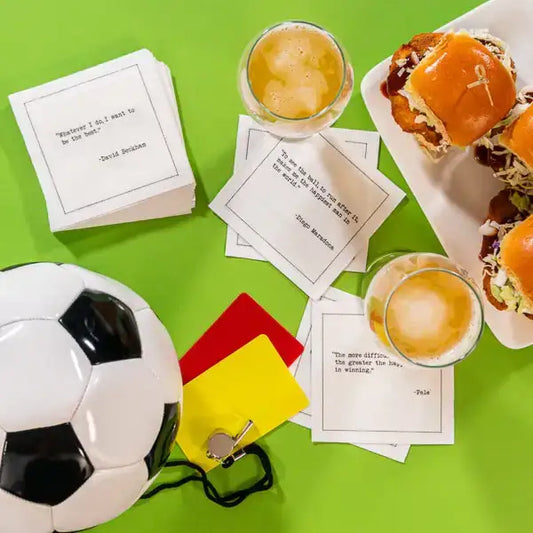 Organic cotton cocktail napkins printed with soccer quotes from David Beckham, Diego Maradona, and Pelé, styled with a soccer ball, referee cards, sliders, and drinks, from Five Star Napkins’ Soccer Legends quote collection.