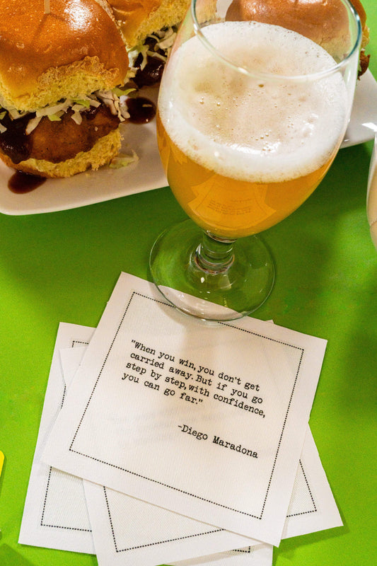 Organic cotton cocktail napkin printed with the quote “When you win, you don’t get carried away. But if you go step by step, with confidence, you can go far,” – Diego Maradona, styled with a beer and sliders, from Five Star Napkins’ Soccer Legends quote collection.