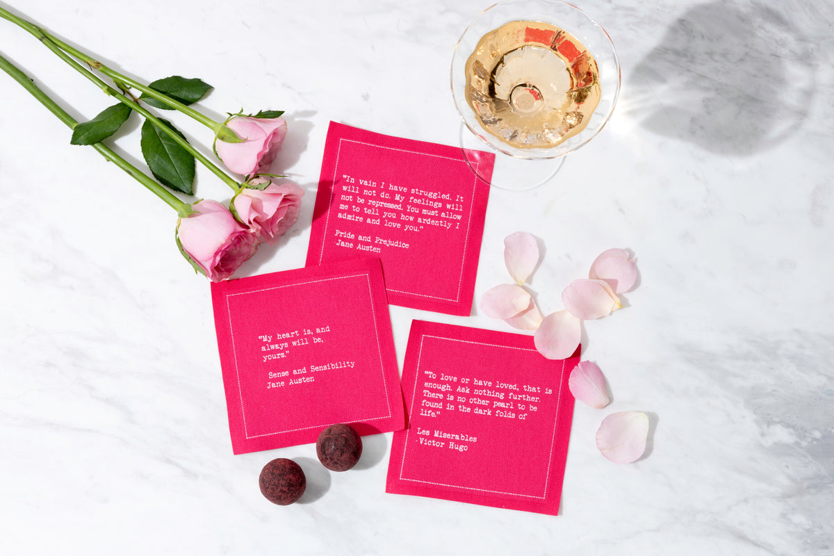 Bright pink Love Quotes cocktail napkins featuring literary passages are arranged with pink roses, rose petals, chocolates, and a glass of rosé on a white marble surface.
