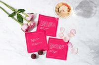 Bright pink Love Quotes cocktail napkins featuring literary passages are arranged with pink roses, rose petals, chocolates, and a glass of rosé on a white marble surface.
