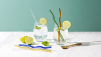 Two iced drinks garnished with lime, styled with wavy and clear glass straws and a gold spoon on a teal and white tabletop.