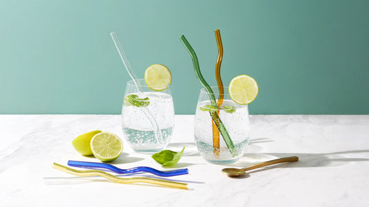 Two iced drinks garnished with lime, styled with wavy and clear glass straws and a gold spoon on a teal and white tabletop.