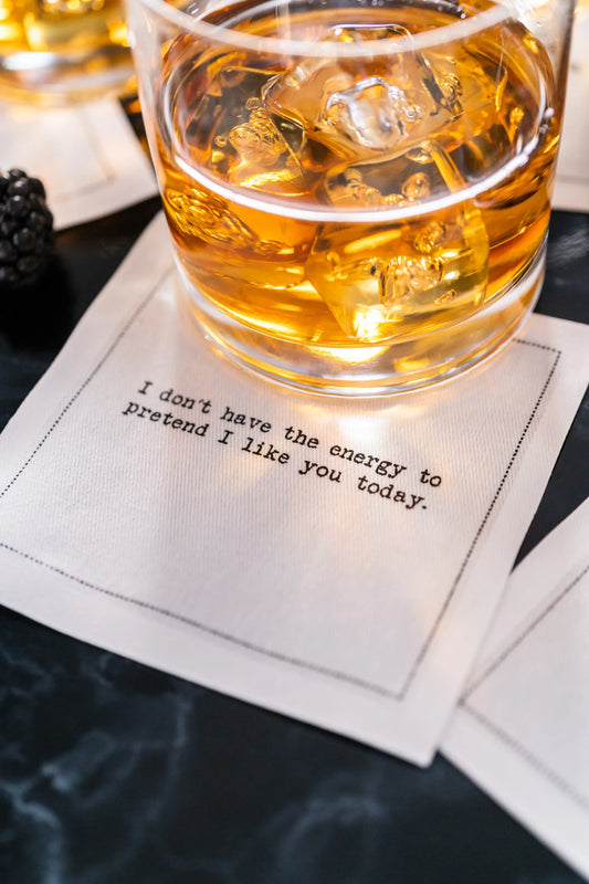 Organic cotton cocktail napkin printed with the quote "I don't have the energy to pretend I like you today," styled under a glass of whiskey on a marble table, from Five Star Napkins' Subversive quote collection