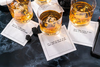 Three organic cotton cocktail napkins styled under whiskey glasses on a marble table, printed with quotes including "Never go to bed angry, stay awake and plot revenge" and "Decaf only works if you throw it at people," from Five Star Napkins' Subversive quote collection
