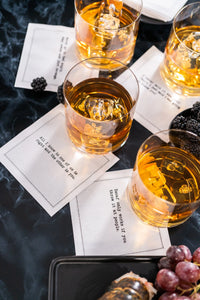 Organic cotton cocktail napkins styled under whiskey glasses on a marble table with berries and grapes, printed with quotes including "All I know is one of us is right and the other is you" and "Decaf only works if you throw it at people," from Five Star Napkins' Subversive quote collection
