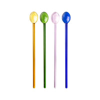 Four glass cocktail swizzle spoons in yellow, green, pink, and blue, standing upright against a white background.