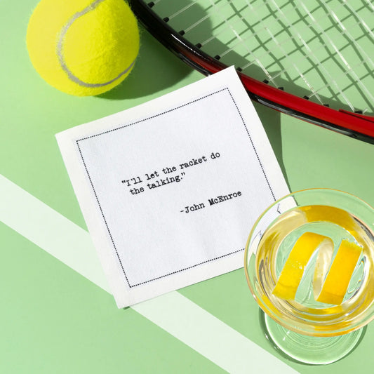 Organic cotton cocktail napkin printed with the quote “I’ll let the racket do the talking,” – John McEnroe, styled with a tennis ball, racket, and citrus cocktail, from Five Star Napkins’ Tennis Legends quote collection.