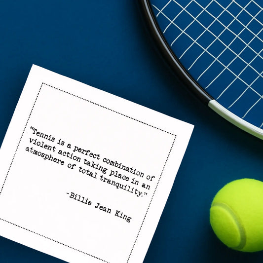Organic cotton cocktail napkin printed with the quote “Tennis is a perfect combination of violent action taking place in an atmosphere of total tranquility,” – Billie Jean King, styled with a tennis racket and ball on a blue court, from Five Star Napkins’ Tennis Legends quote collection.