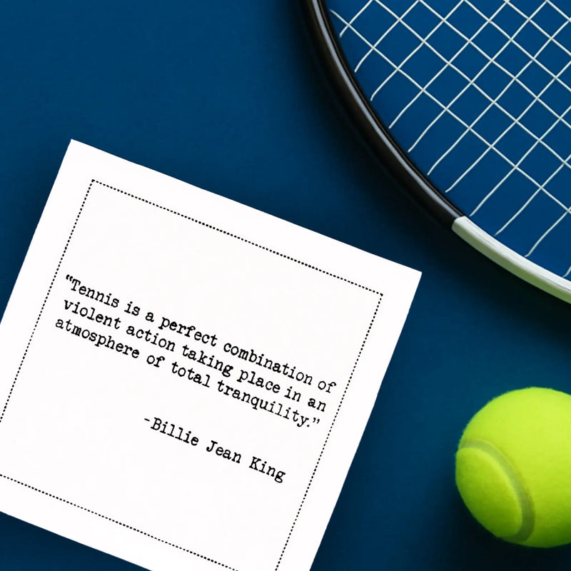 Organic cotton cocktail napkin printed with the quote “Tennis is a perfect combination of violent action taking place in an atmosphere of total tranquility,” – Billie Jean King, styled with a tennis racket and ball on a blue court, from Five Star Napkins’ Tennis Legends quote collection.