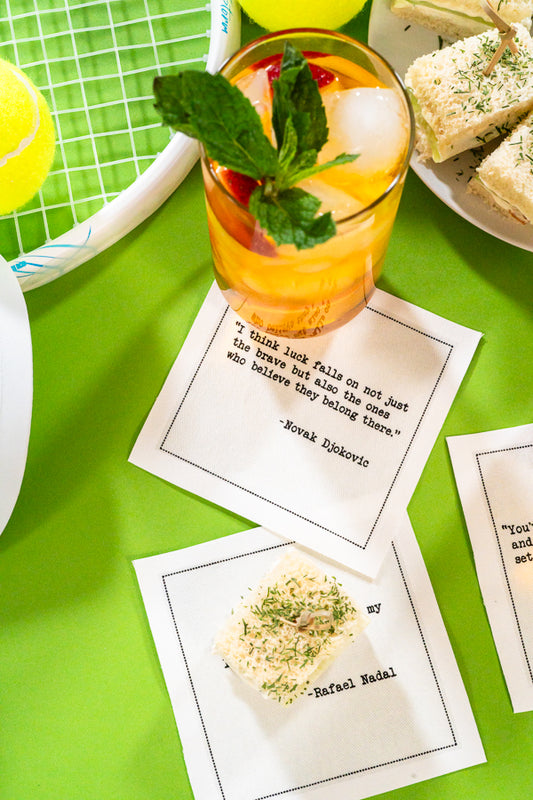 Luxury cocktail napkins with tennis quotes, tea sandwiches, and drink on green table with racket