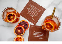 Two Persimmon organic cotton cocktail napkins with printed Thanksgiving quotes, styled with drinks and blood orange slices on a marble table.
