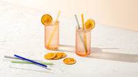 Two glasses of iced citrus drinks garnished with dried orange slices, styled with colorful glass straws and a cleaning brush on a sunlit tabletop.